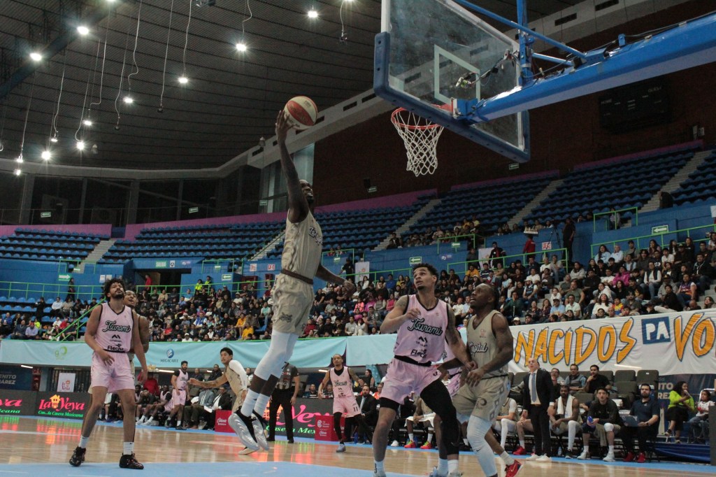 An intense basketball game in progress, featuring players from opposing teams lunging for a slam dunk, with a crowd watching in a well-lit arena.