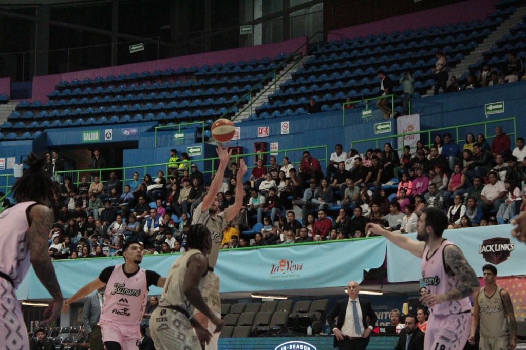 A basketball player jumps to shoot the ball during a game, with spectators in the background watching intently.