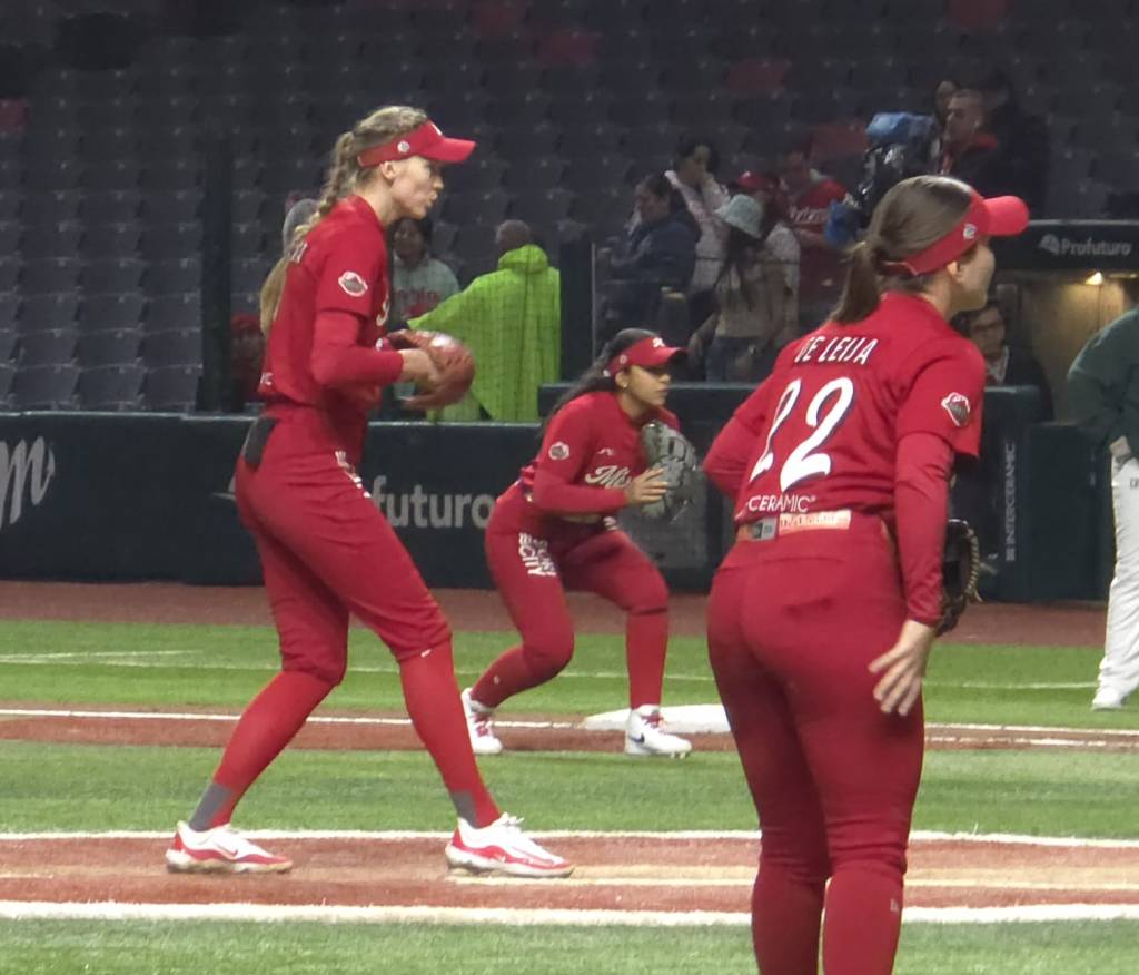A baseball pitcher in a red uniform prepares to throw the ball, with two teammates in the background also in red uniforms. The scene takes place on a baseball field, with empty stadium seating visible in the background.
