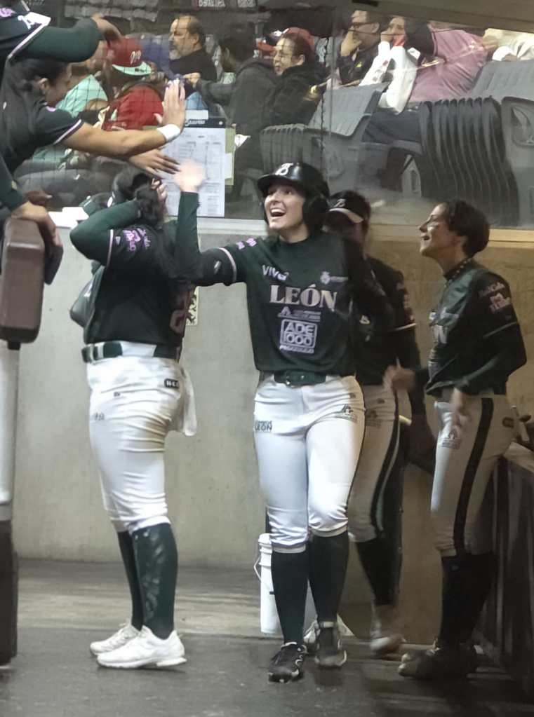 A group of female softball players celebrating in the dugout, with one player smiling and high-fiving her teammates. The players are dressed in matching uniforms, and spectators can be seen in the background.